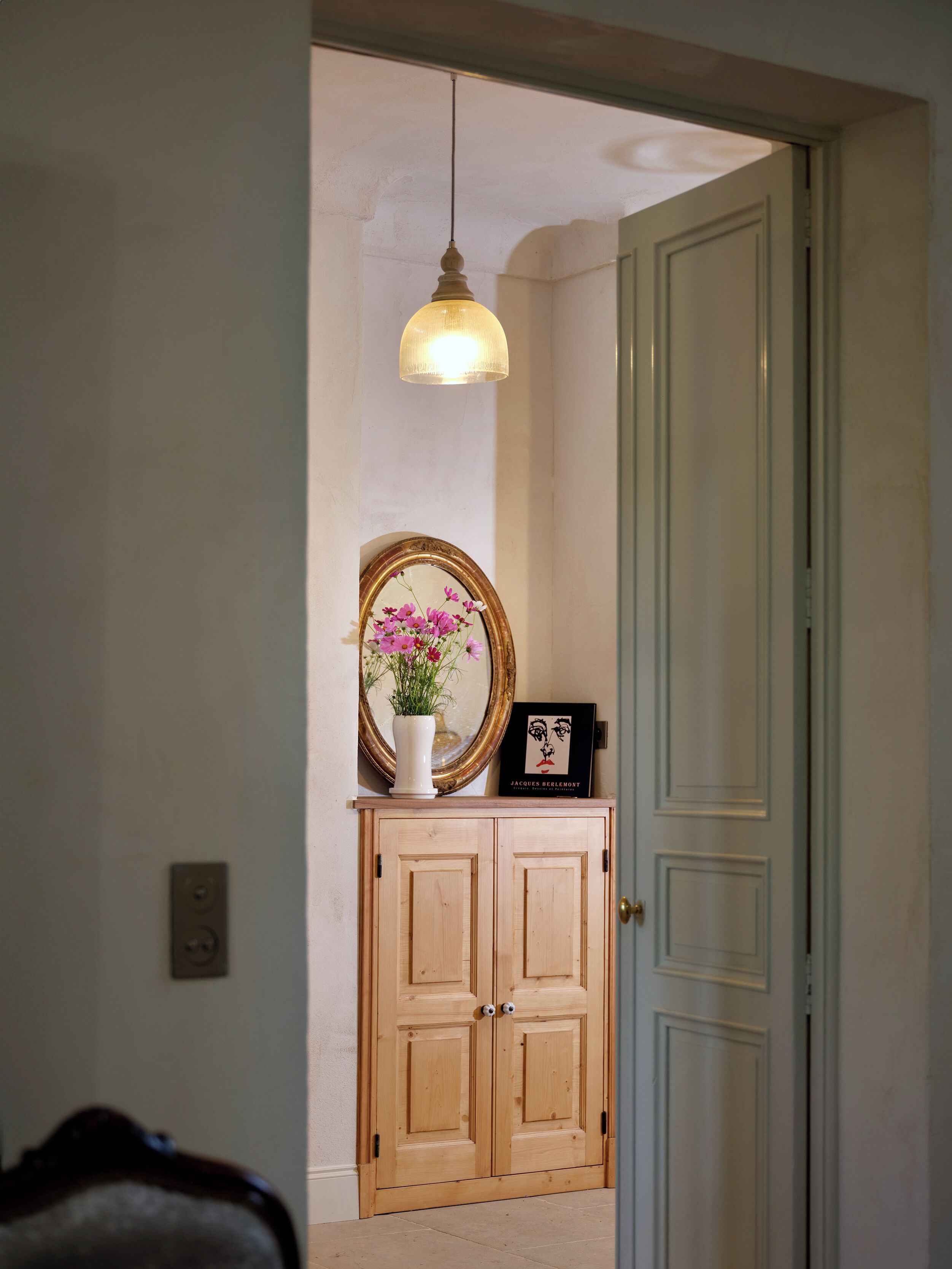 A hallway with a wooden double door, a white vase with pink flowers, a framed mirror, and a pendant light.