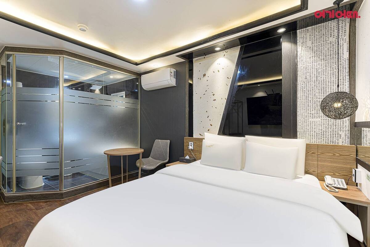 A modern hotel room with a large bed, a glass-enclosed walk-in closet, a wooden nightstand, and a large window with decorative curtains.