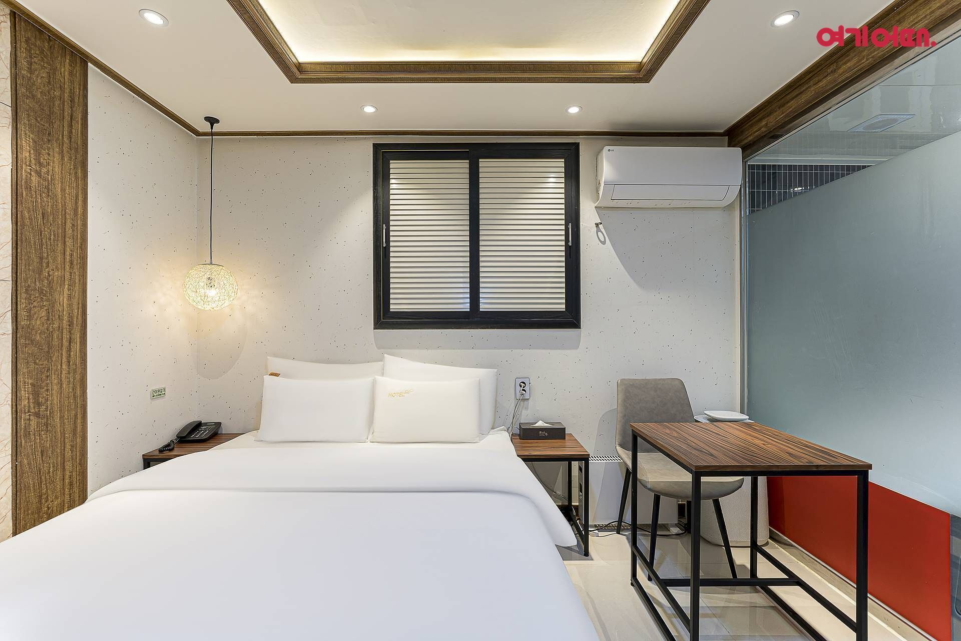 Une chambre d’hôtel comprenant un lit, une table de chevet, une lampe, une fenêtre, un appareil de climatisation et un bureau avec une chaise.