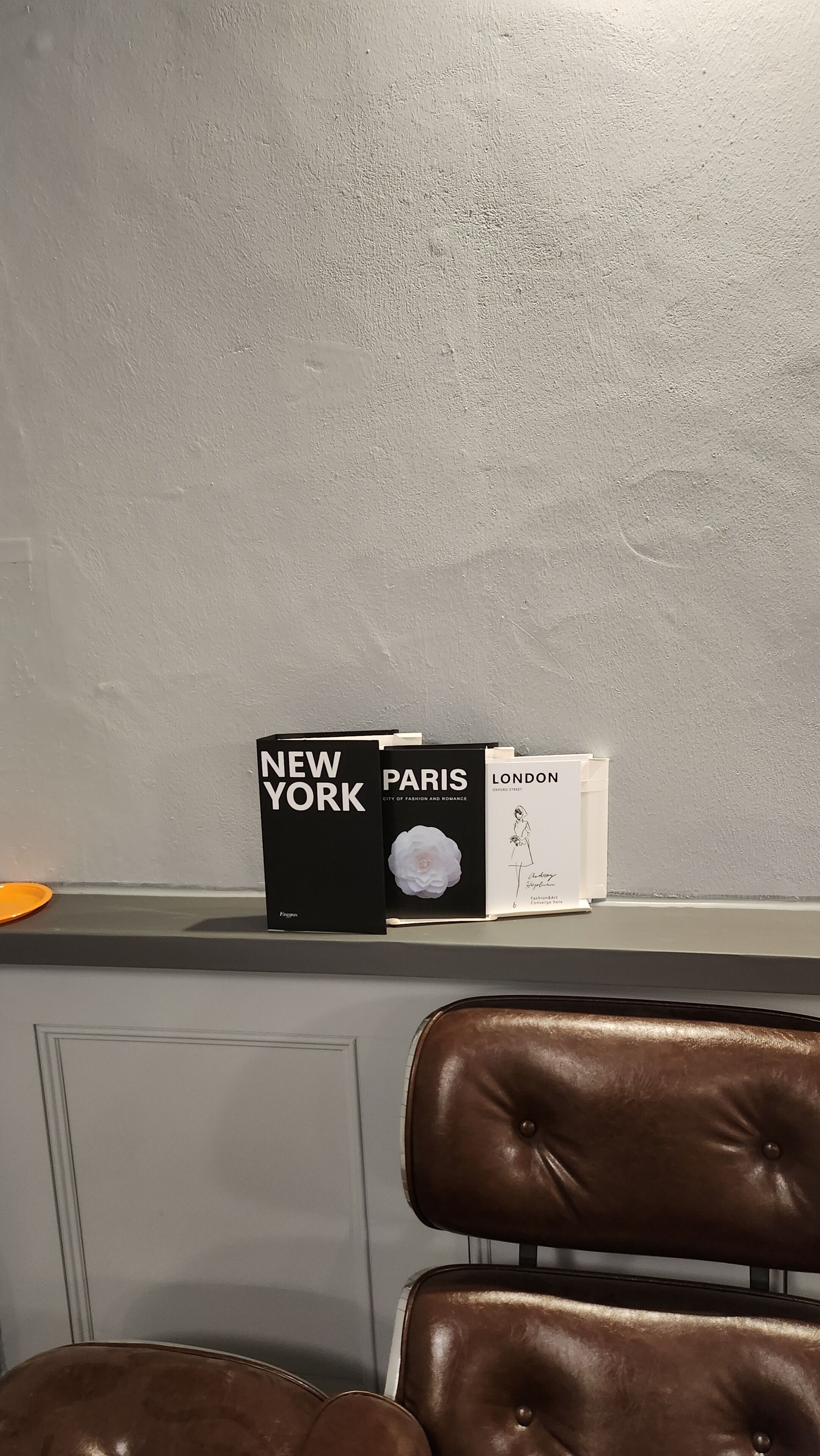 A shelf with books titled 'New York,' 'Paris,' and 'London,' and a white plate with a flower.