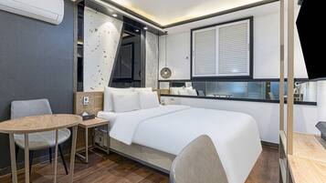 A compact hotel room with a bed, a small table, a chair, and a window with blinds.