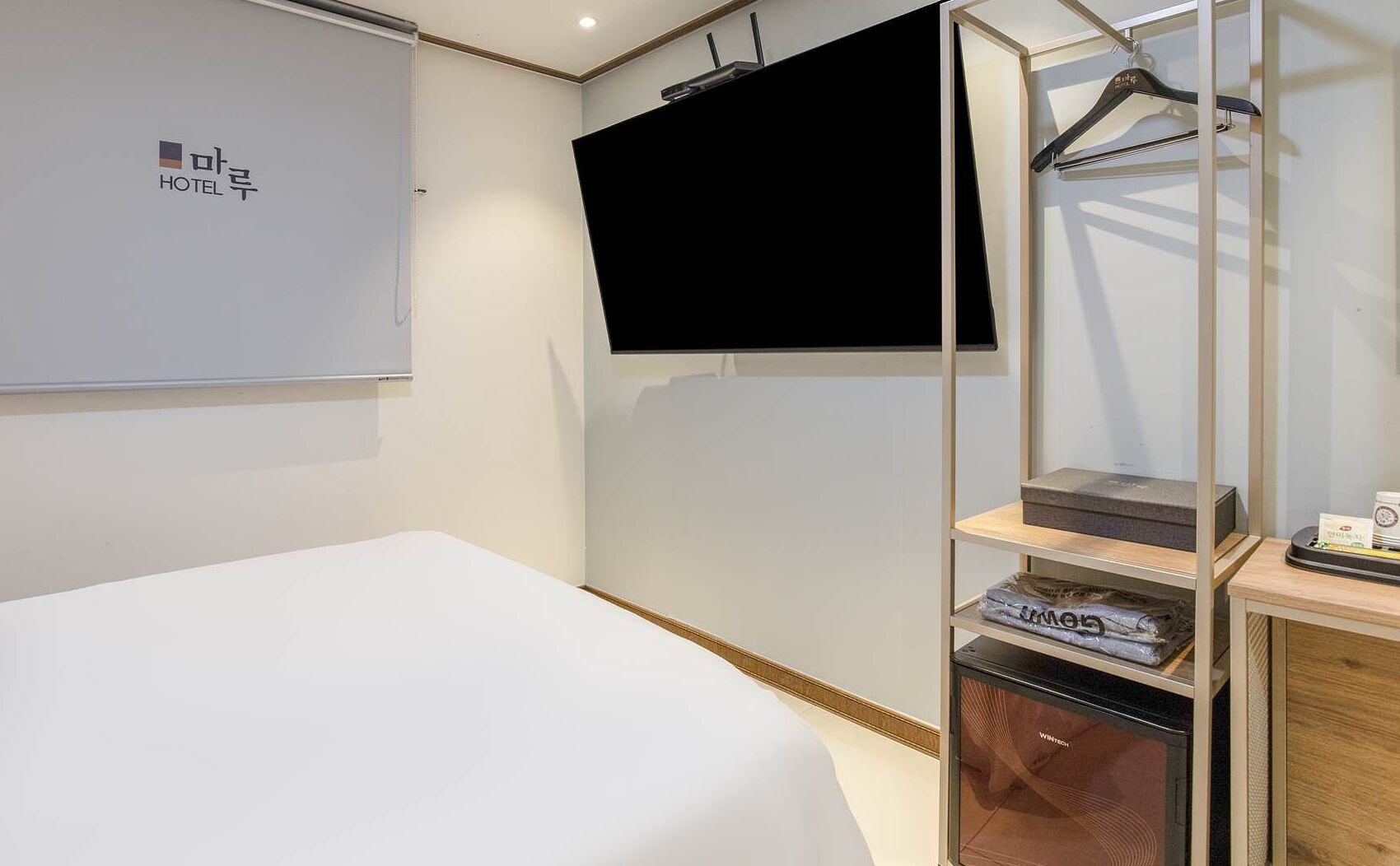 A hotel room with a bed, a TV mounted on the wall, a wardrobe with shelves, and a small table with a cup.