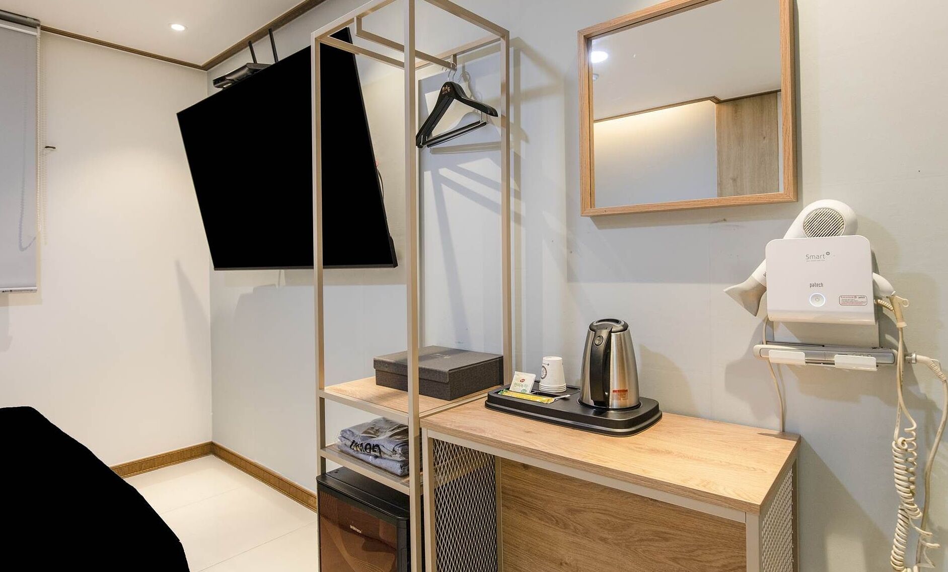 Una habitación de hotel moderna con un tocador de madera, un secador de pelo, una tetera y un televisor montado en la pared.