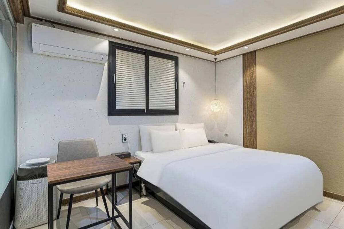 A hotel room with a bed, a desk, a chair, a window with blinds, and a wall-mounted air conditioner.