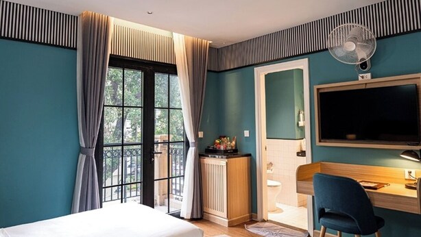 A hotel room with a bed, a desk, a chair, a television, a fan, and a view of the outdoors.