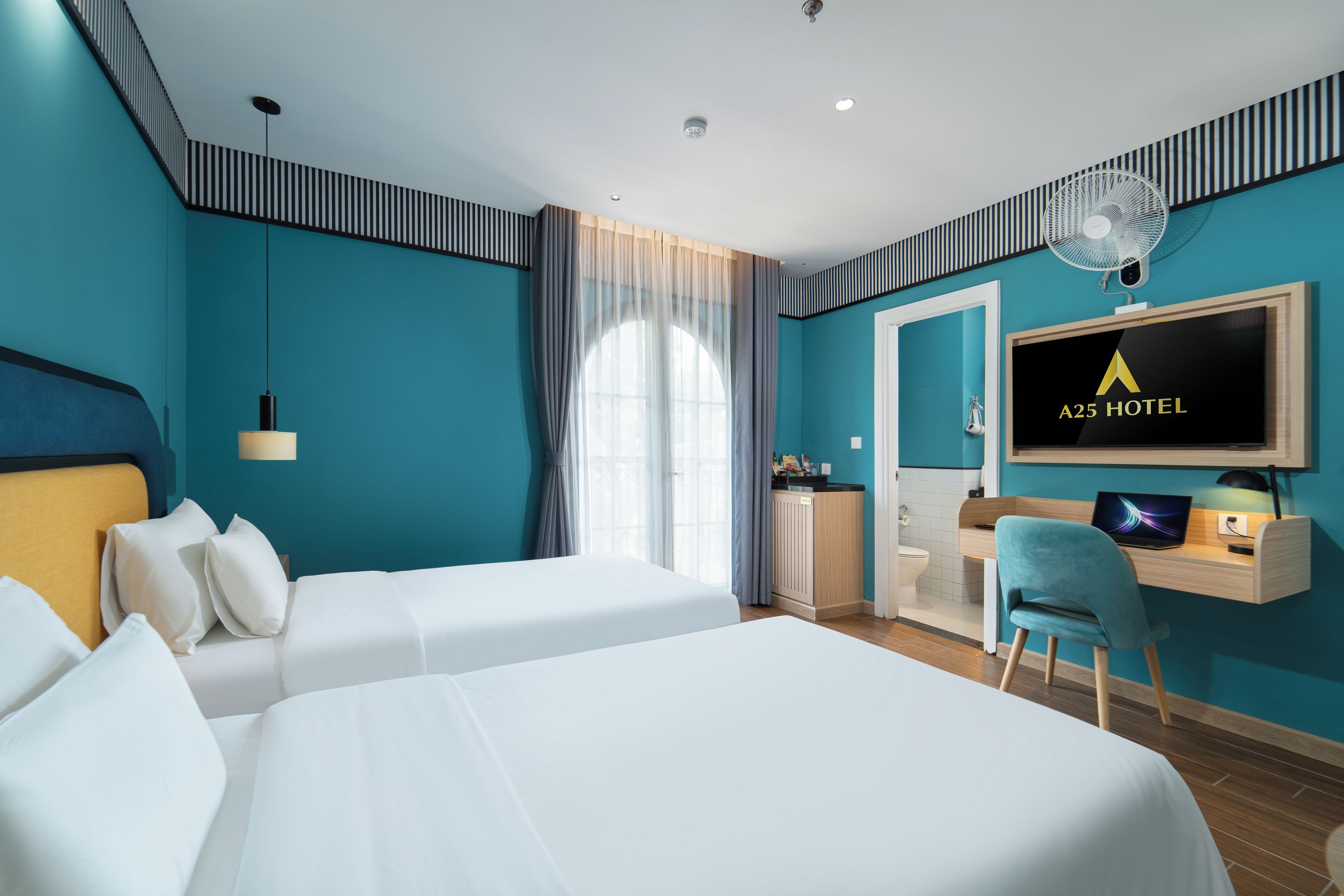 Habitación de hotel con dos camas, un escritorio con computadora, televisor y ventilador.