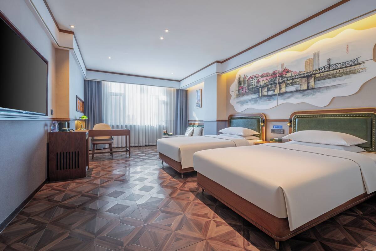 Habitación de hotel con dos camas, un escritorio, una silla y un amplio ventanal con cortinas.