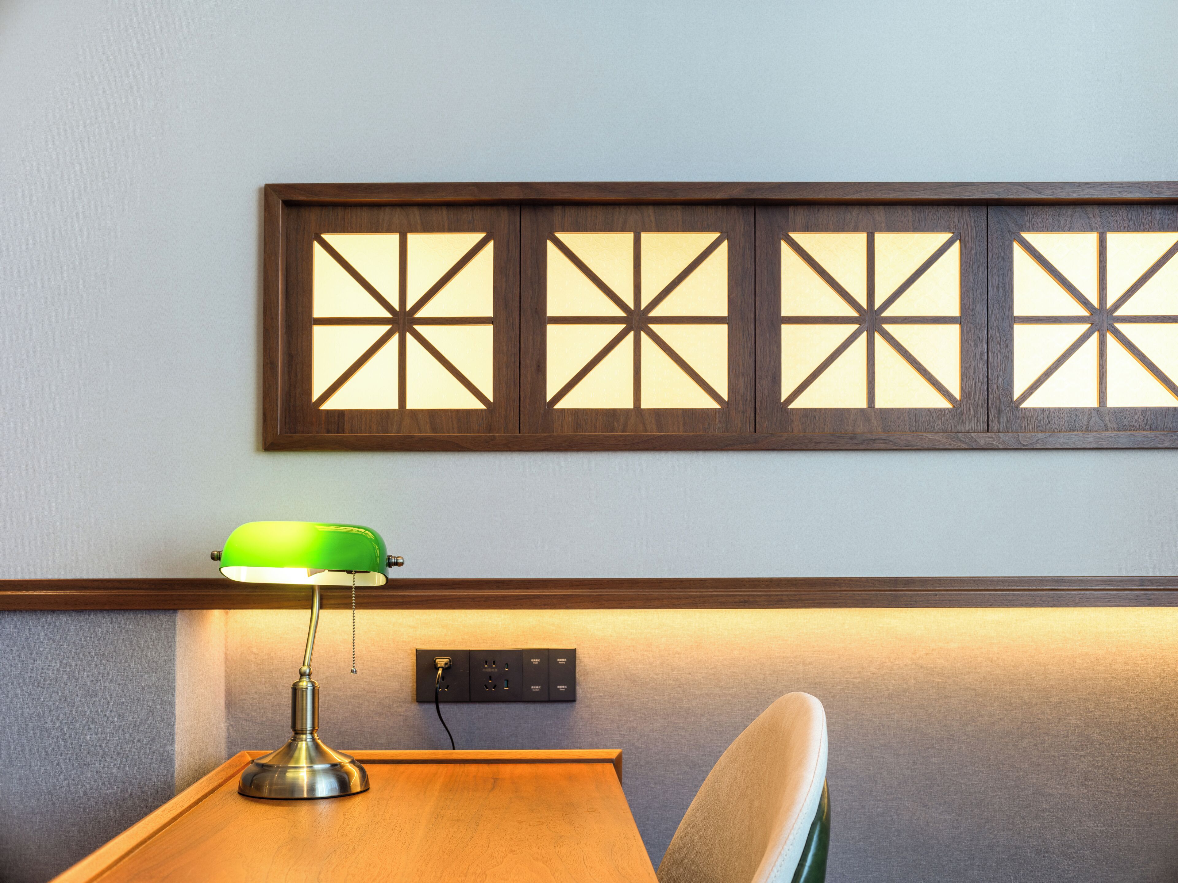 A wooden desk with a green desk lamp, a power outlet, and a wall-mounted panel with geometric cutouts.