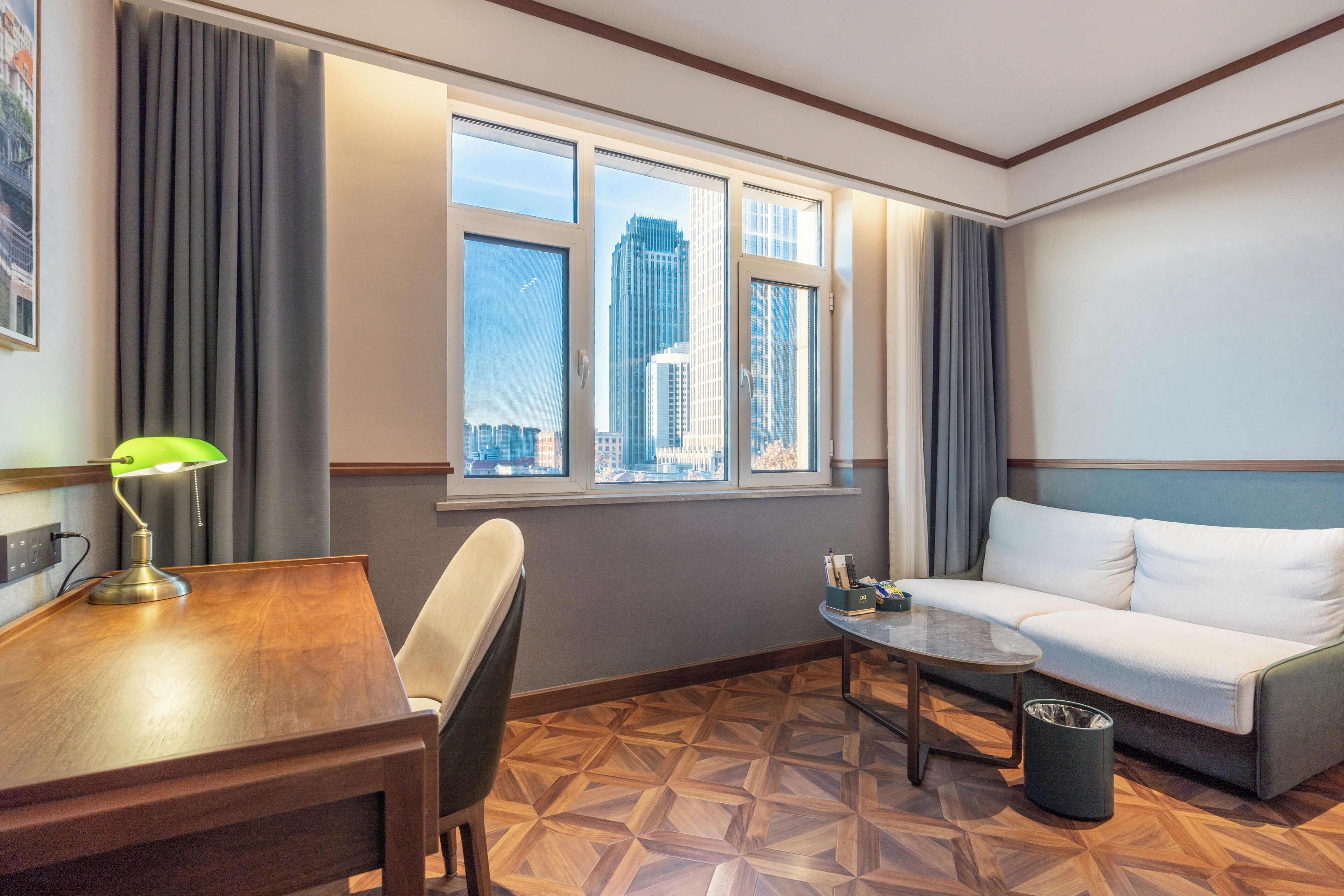 A hotel room with a wooden desk, a green desk lamp, a white sofa, a small round table, and a view of tall buildings through large windows.