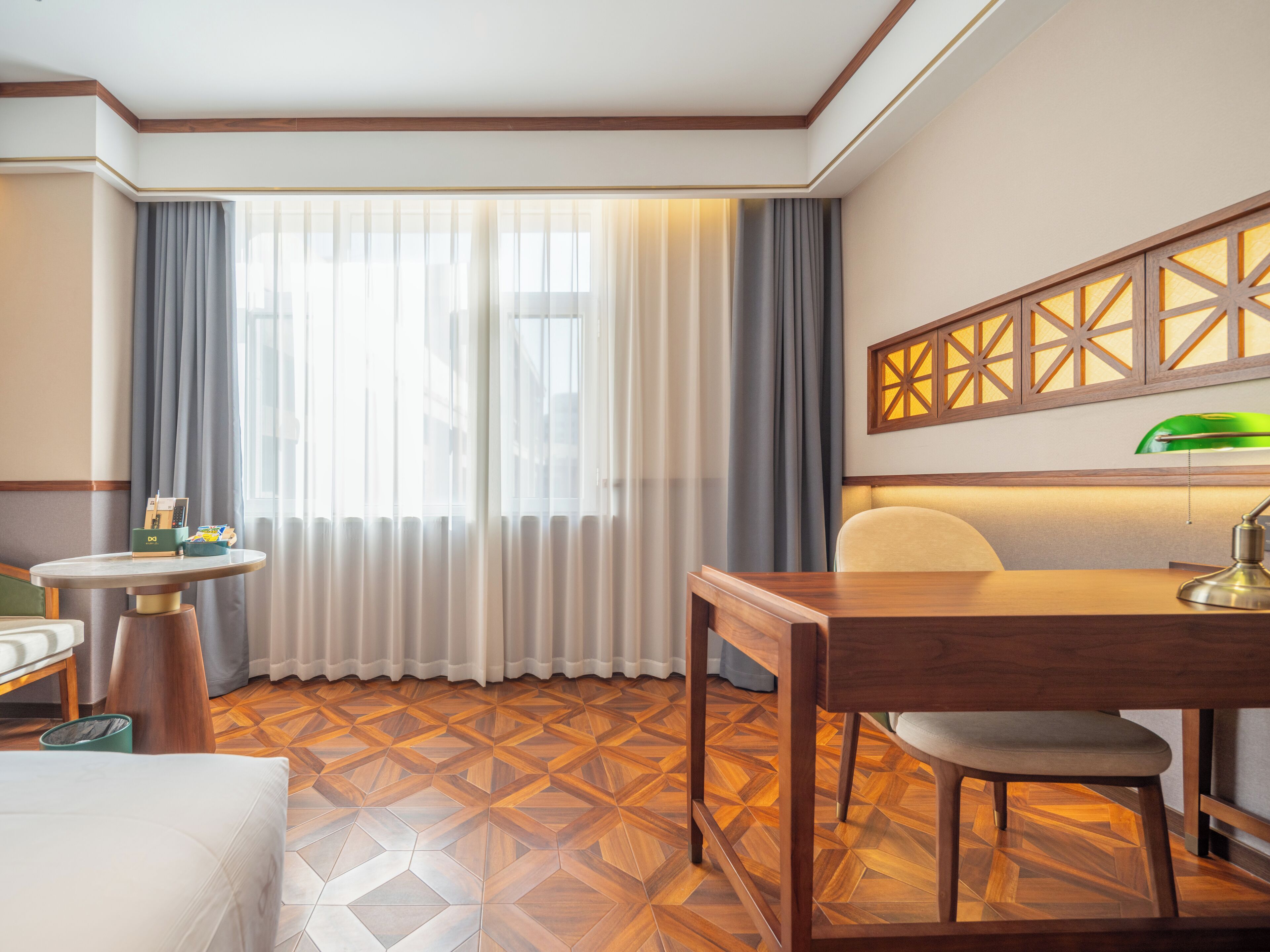 Una habitación de hotel con una mesa de comedor de madera, una silla, una mesita redonda con una lámpara y una cama con cabecera blanca.
