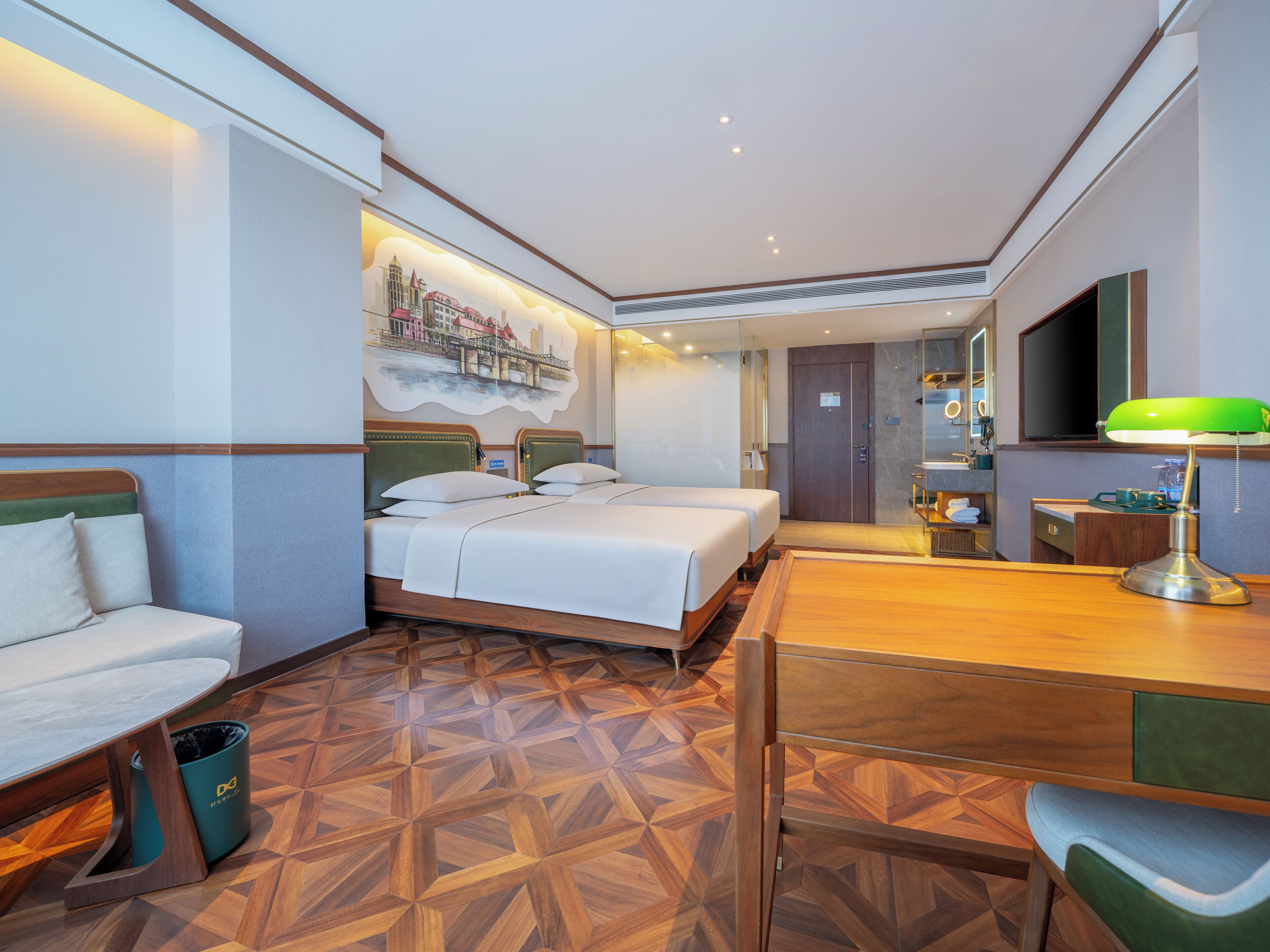 Habitación de hotel con dos camas, un escritorio, una silla, un cubo de basura y un baño visible a través de una puerta de cristal.