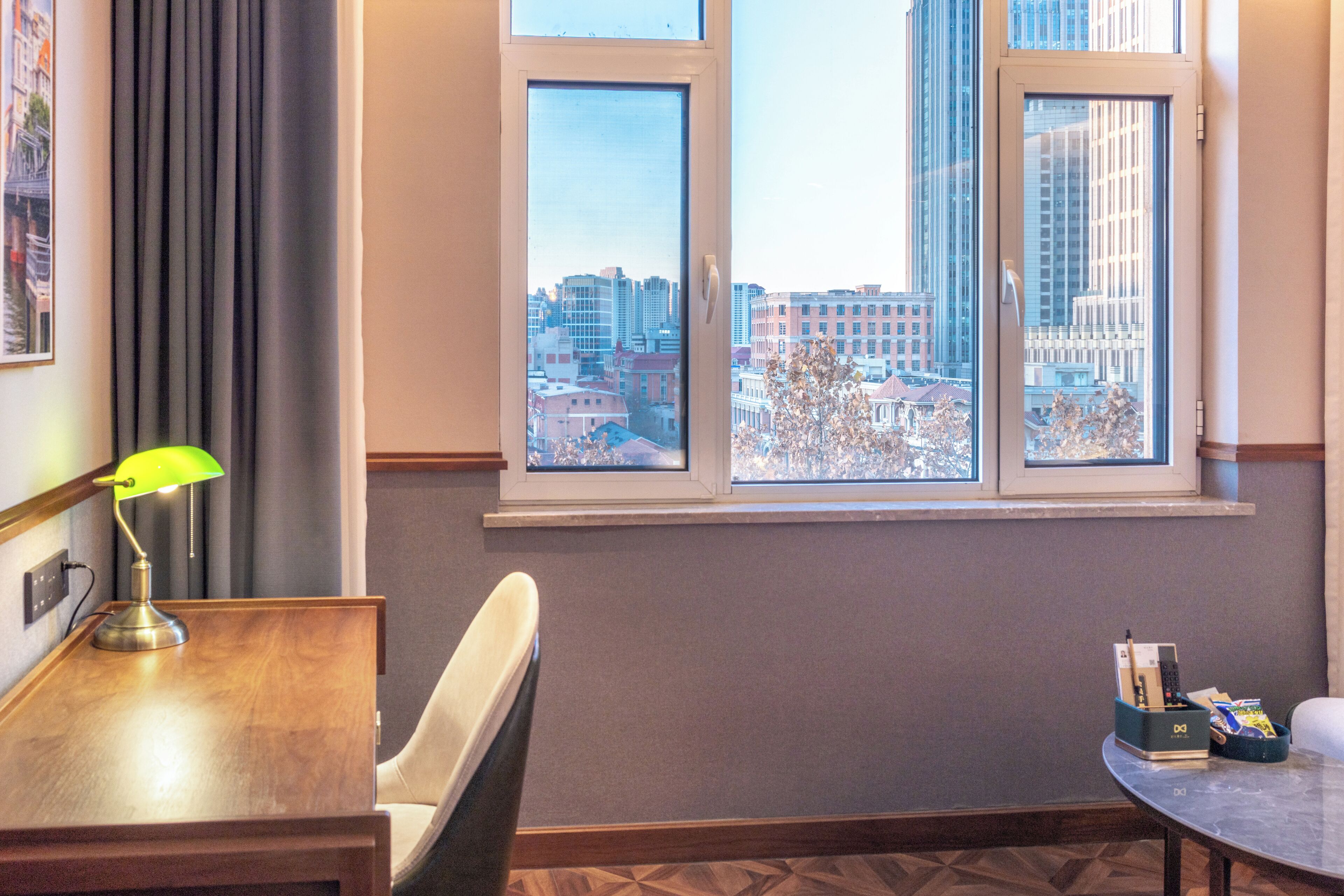 A hotel room with a city view, a desk with a lamp, a chair, and a small table with a box of toiletries.