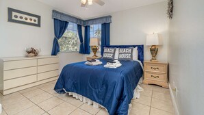 Quarto com cama, cômoda, mesa de cabeceira, ventilador de teto e janela com cortinas.