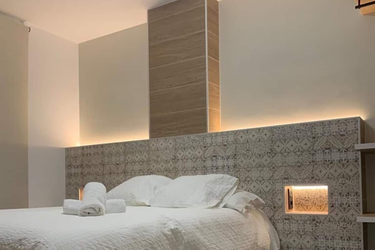 Une chambre à coucher moderne avec un lit, une tête de lit ornée d’un motif décoratif et une petite étagère éclairée.