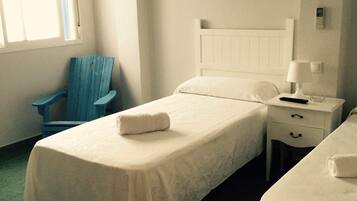 Une chambre simple et bien rangée, avec deux lits simples, une petite table avec une lampe, une chaise et une fenêtre avec des stores.