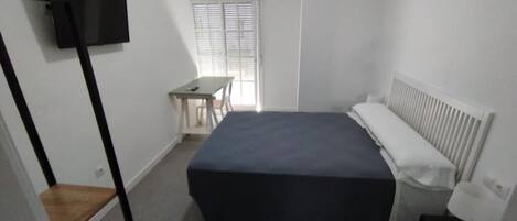 A small, well-lit room with a bed, a desk, and a TV mounted on the wall.