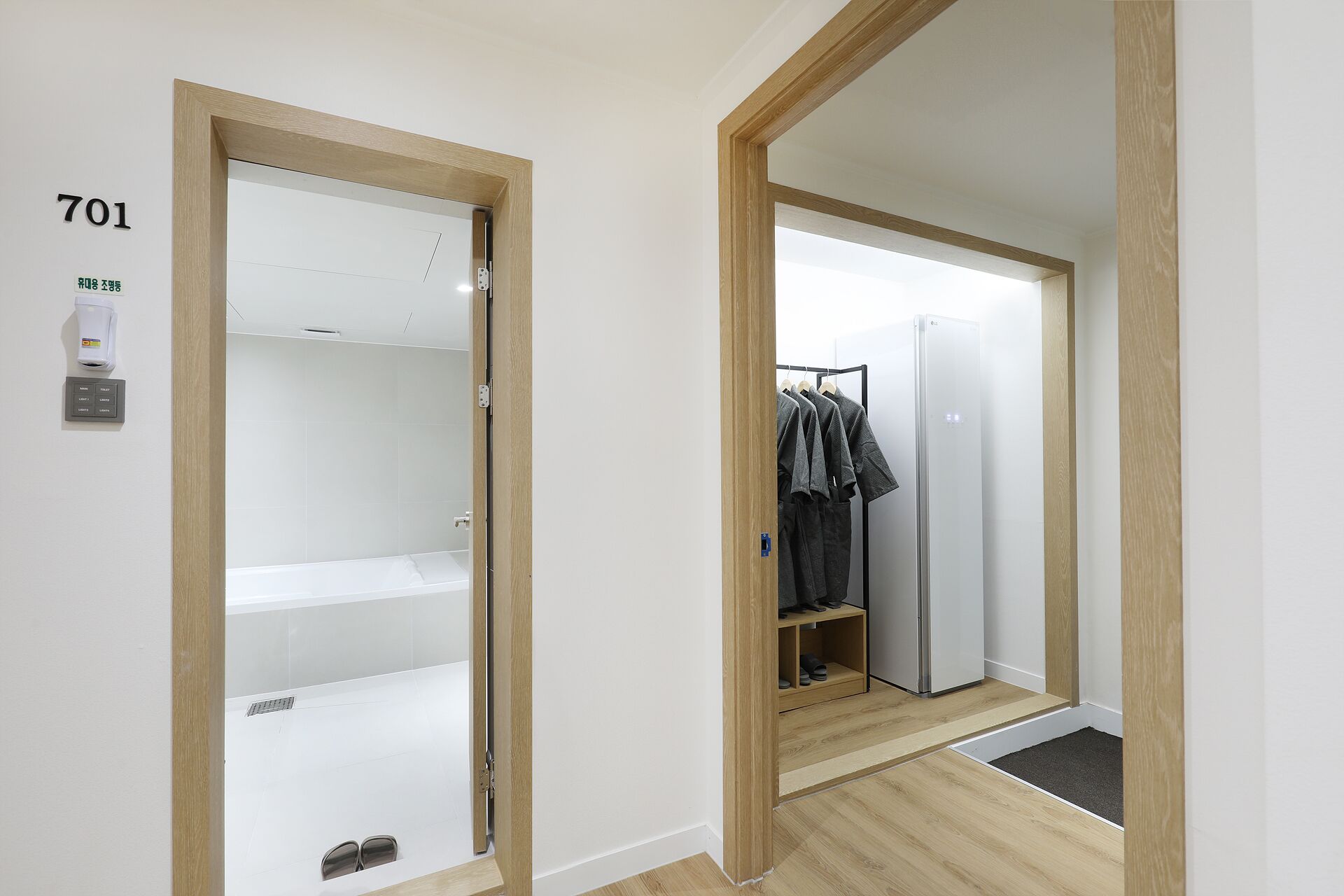 Habitación de hotel con puerta de madera, pared blanca con señal de número, dispensador de toallas, panel de control, zona de ducha, perchero y refrigerador.