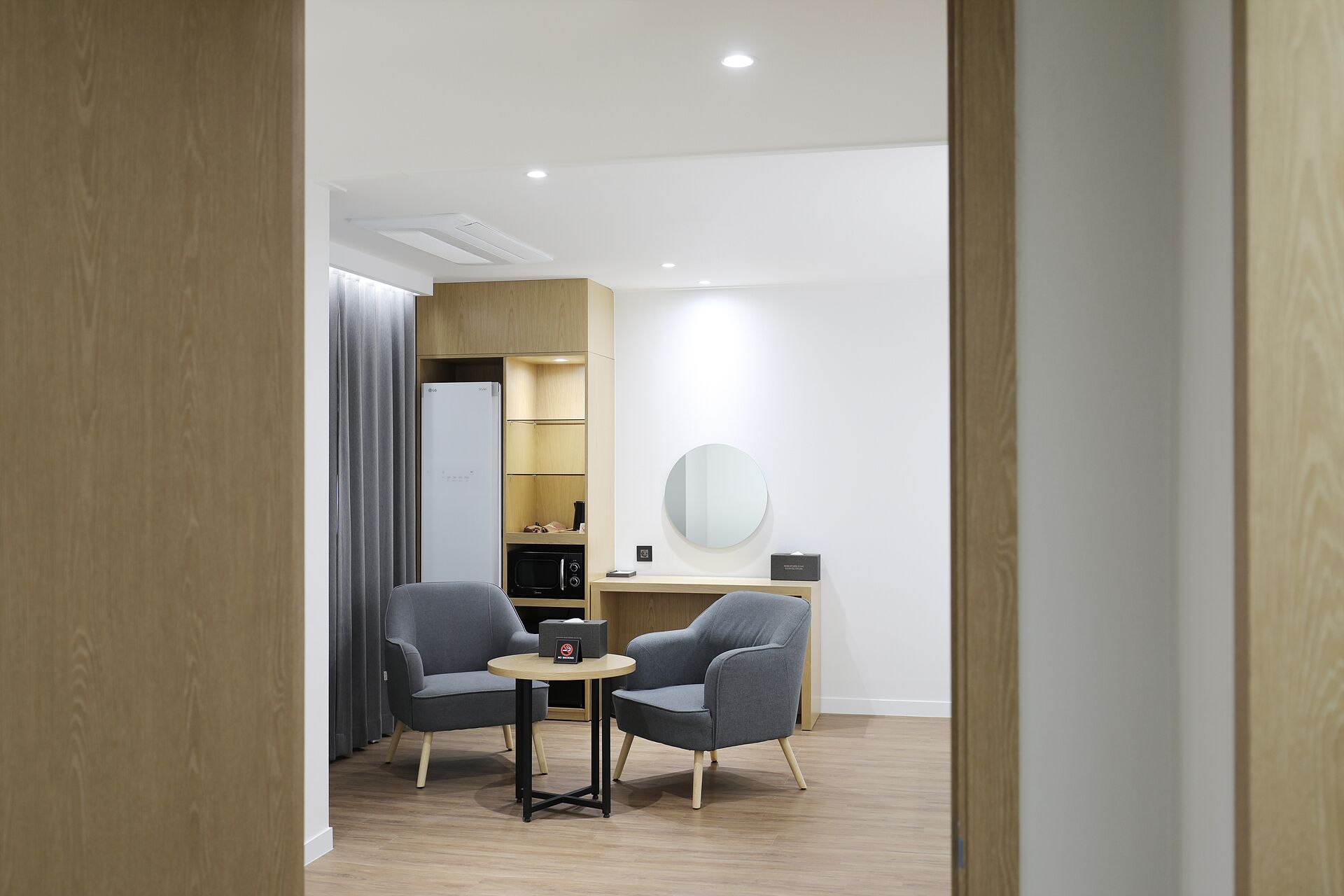Habitación de hotel moderna con suelo de madera, dos sillones grises, una mesita redonda, armario empotrado, microondas y un espejo redondo en la pared.