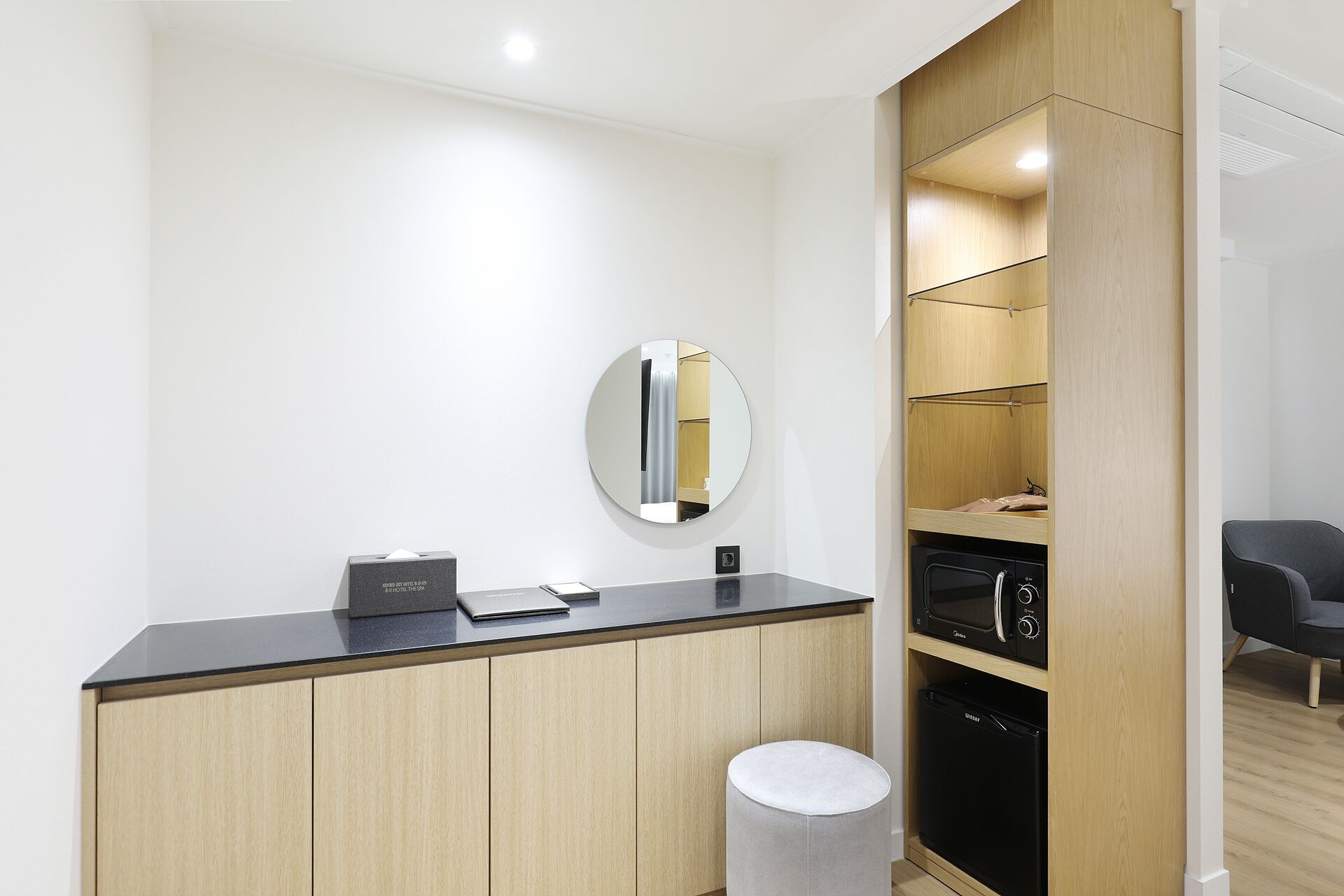 A modern hotel room with a wooden cabinet, a round mirror, a microwave, and a compact refrigerator.