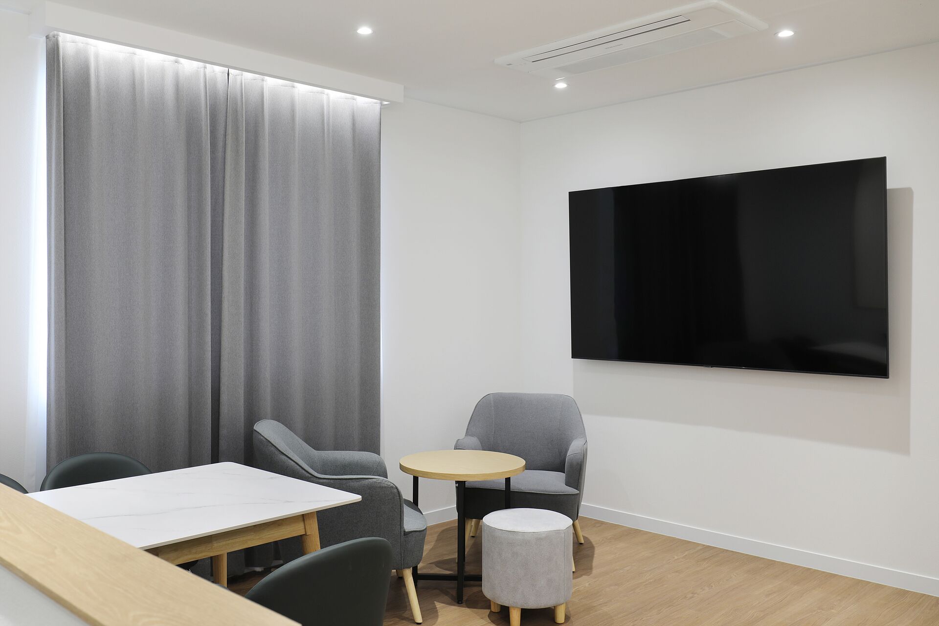 A modern room with a large flat-screen TV mounted on the wall, a grey sofa, a small round table, and a wooden desk with chairs.