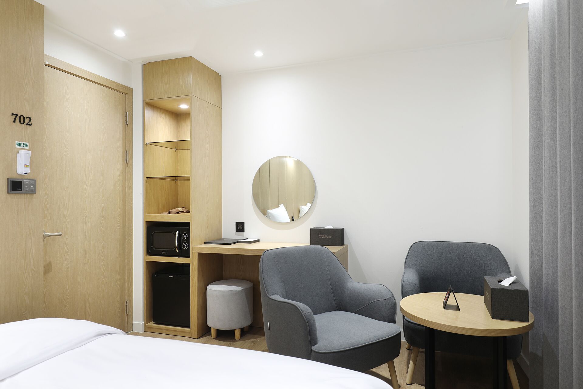 Ein Hotelzimmer mit einem Bett, einem Schreibtisch, zwei Stühlen, einem runden Spiegel, einem kleinen Tisch, einem Mülleimer und einem Kleiderschrank.