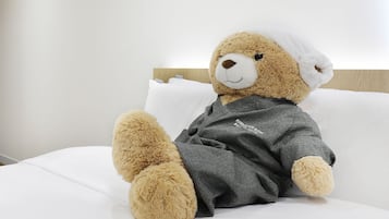 A plush bear in a grey robe sitting on a white bed with a wooden headboard.