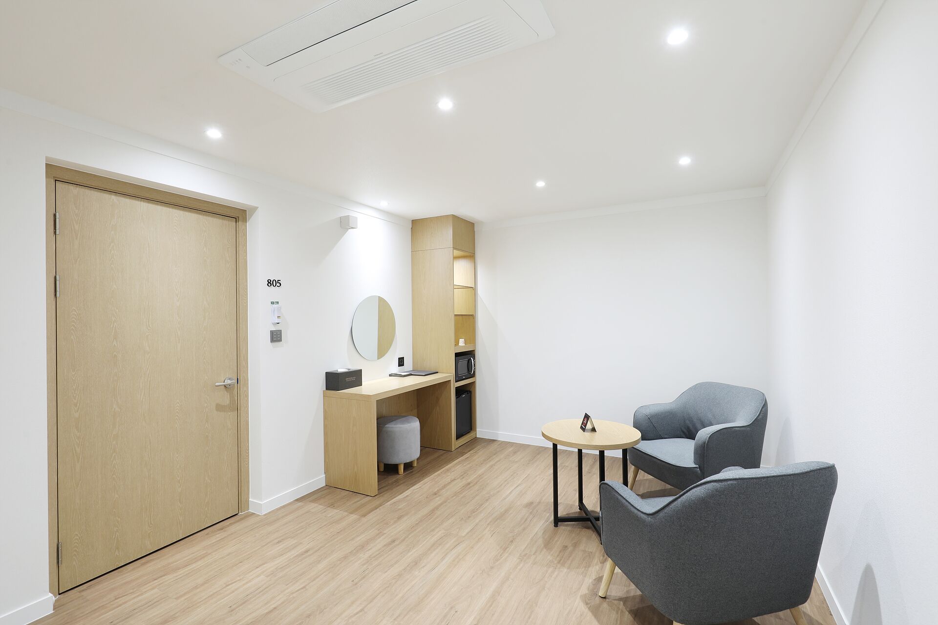Une chambre moderne avec une porte en bois, un bureau doté d’un miroir, une petite table ronde et deux chaises grises.