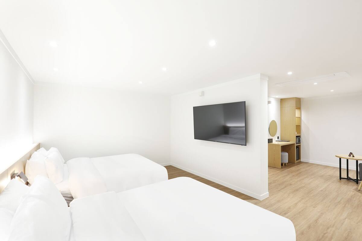 A modern hotel room with two beds, a flat-screen TV mounted on the wall, and a small kitchenette area.