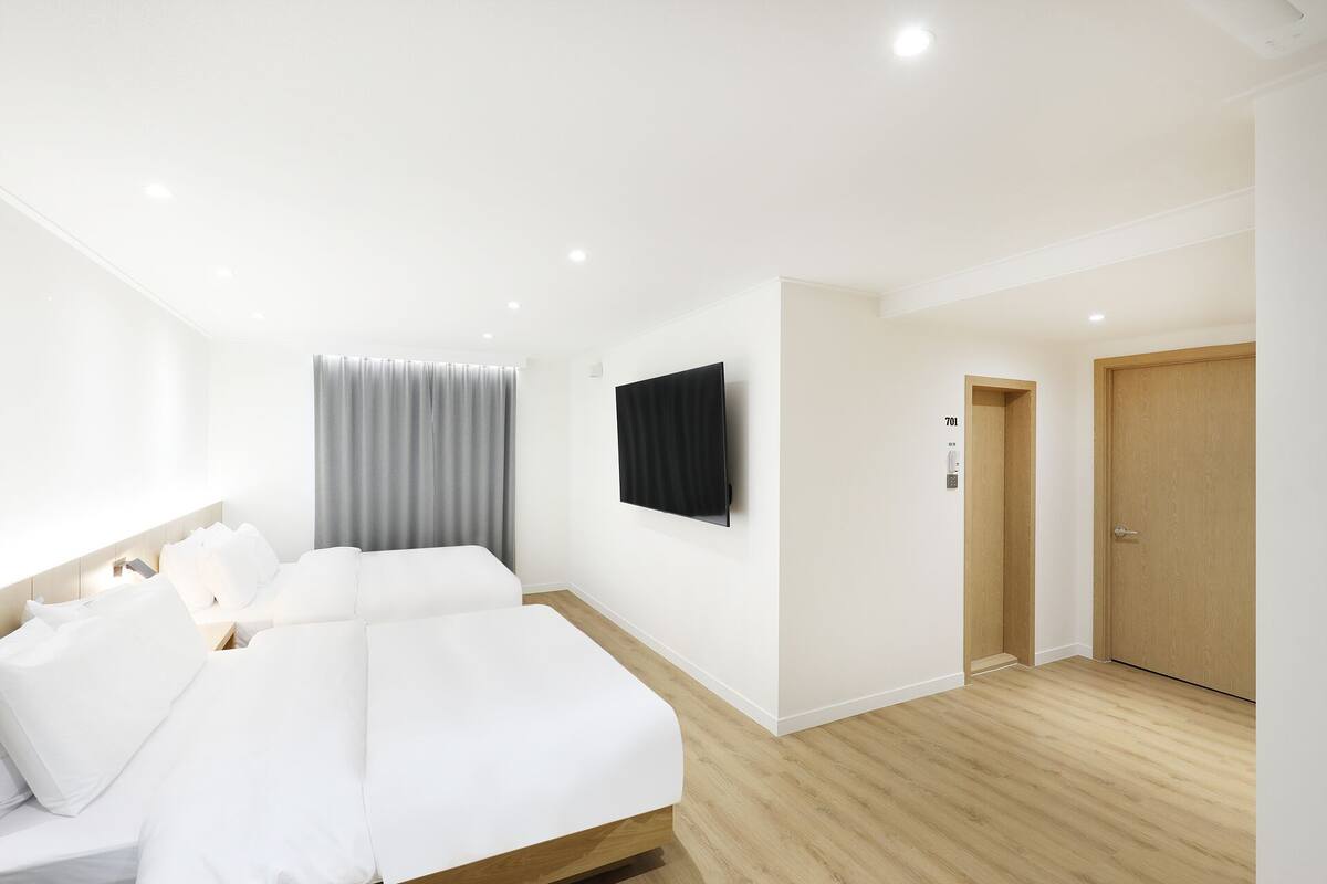 A hotel room with two beds, a flat-screen TV, a wooden door, and a light-colored floor.