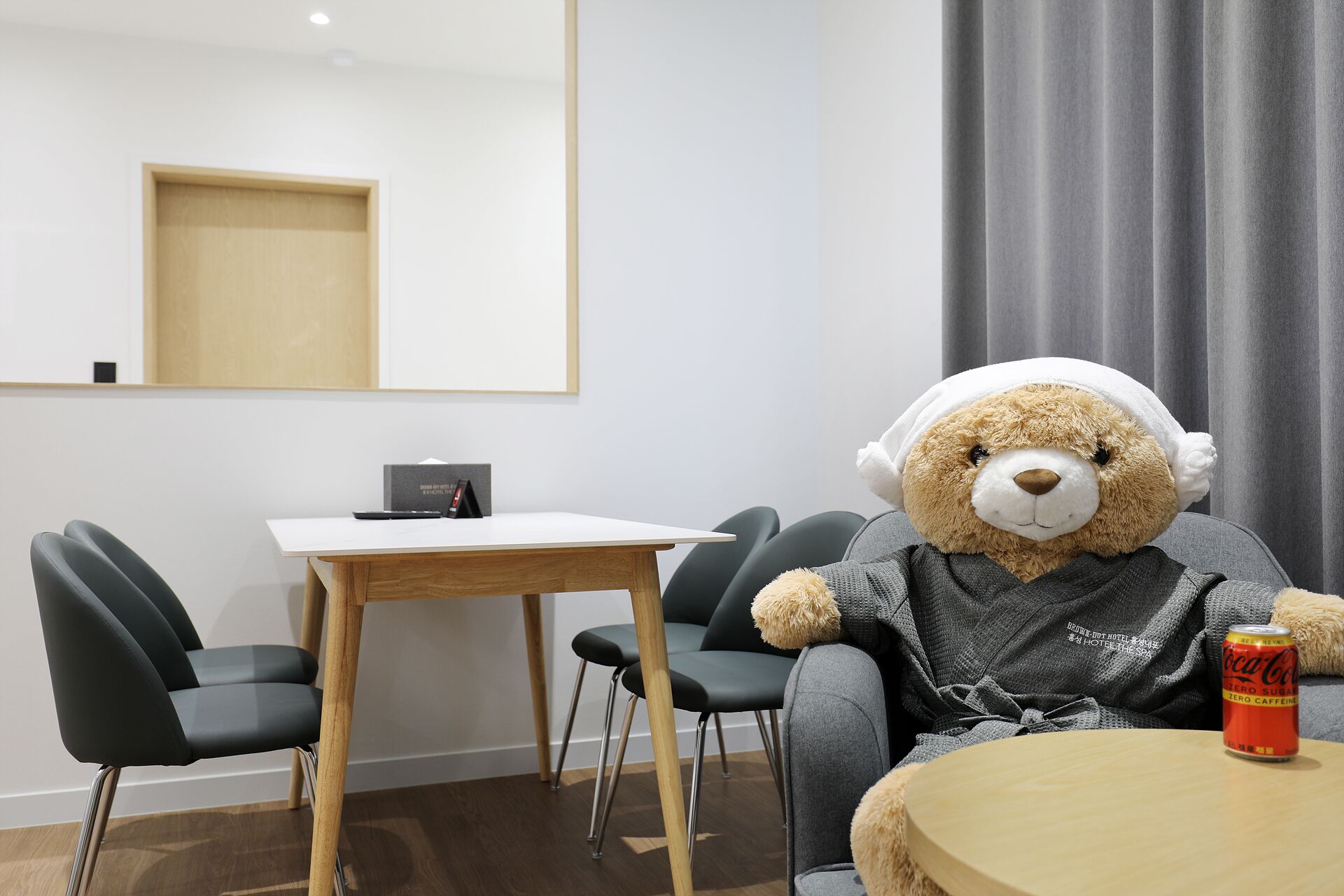 Une pièce avec une table en bois, des chaises noires, un grand ours en peluche portant un casque audio et une canette de Coca-Cola posée sur une table ronde.