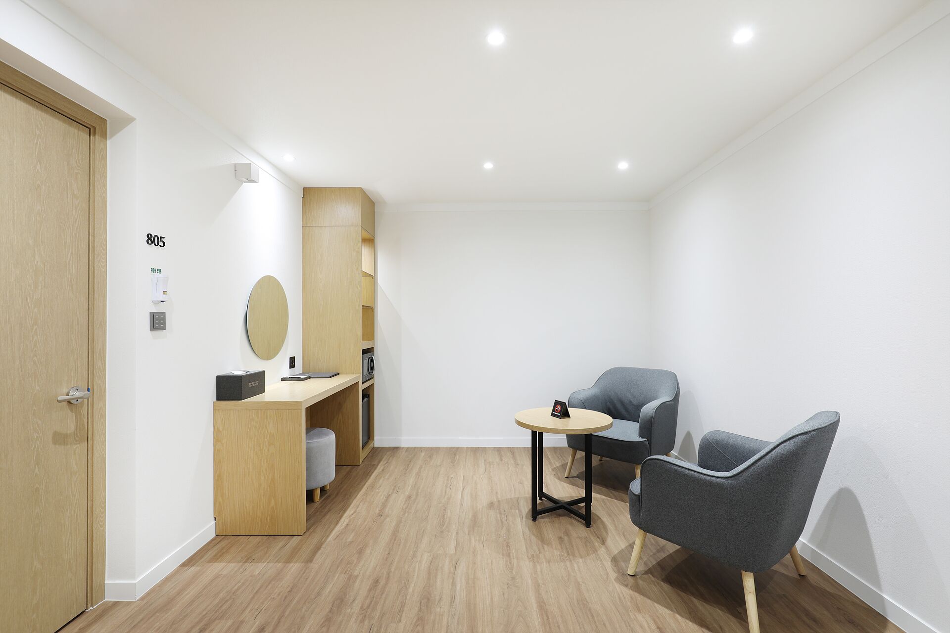 A modern hotel room with a wooden floor, a desk with a mirror, two grey armchairs, and a small round table.