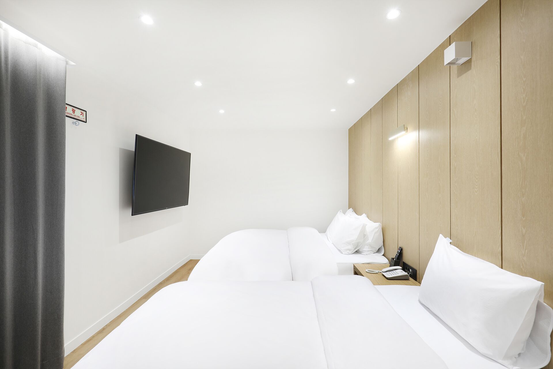A hotel room with two beds, a flat-screen TV mounted on the wall, a wooden panel wall, and a bedside table with a phone.