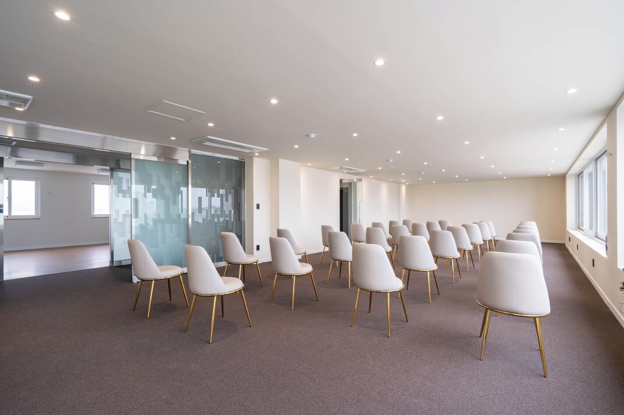 A conference room with rows of white chairs, a glass partition, and a large window.