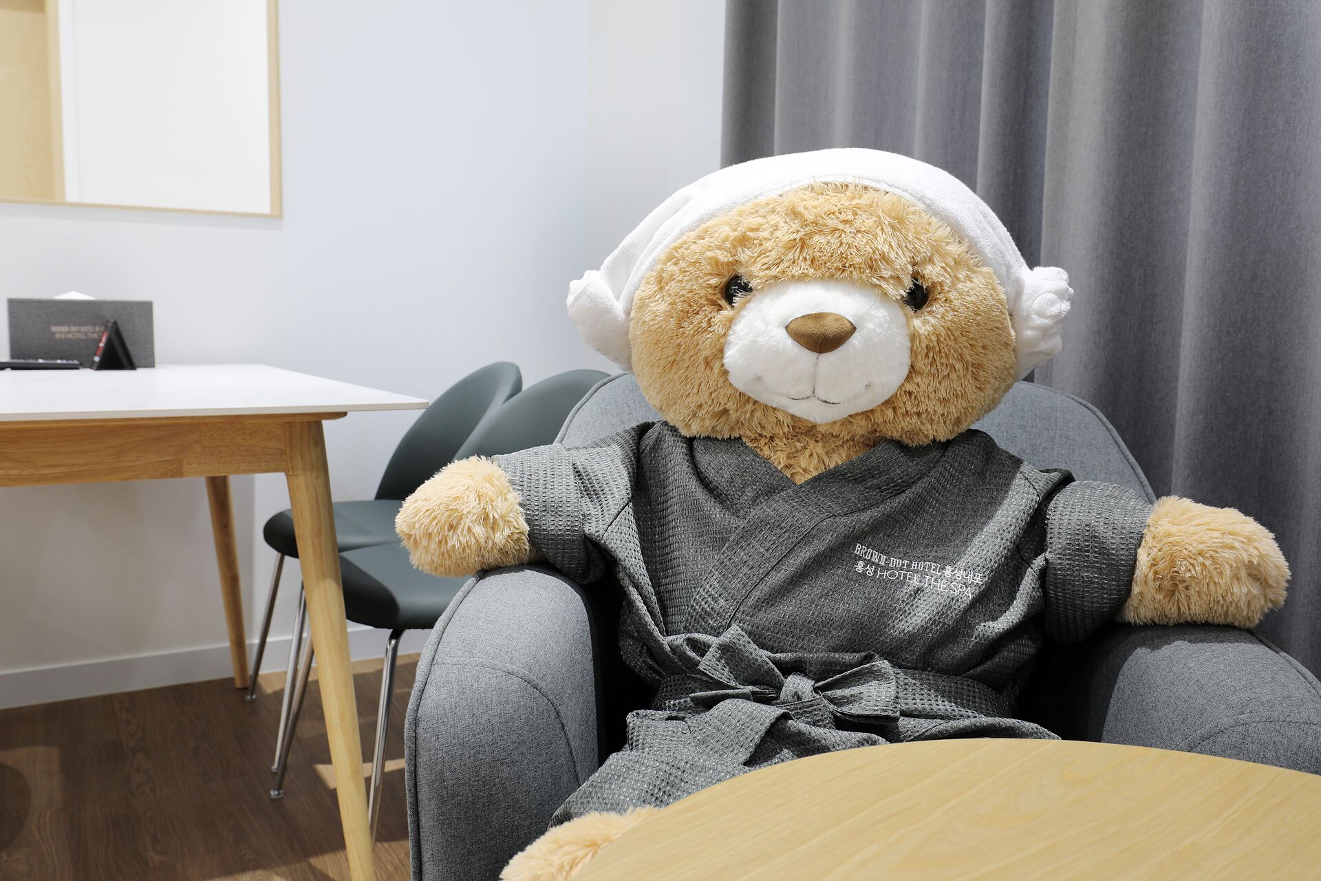 A plush bear wearing headphones, seated at a table in a room with a grey chair, a wooden table, and a grey curtain in the background.