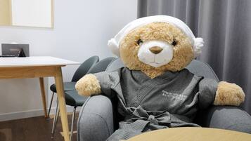 A plush bear wearing headphones, seated at a table in a room with a grey chair, a wooden table, and a grey curtain in the background.