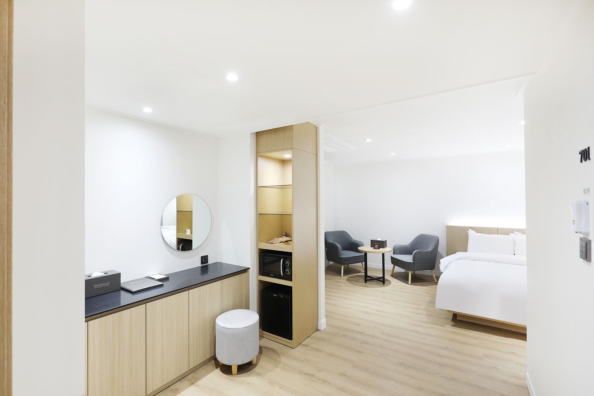 A modern hotel room with a wooden floor, a bed, a desk with a mirror, a small round stool, and two chairs.