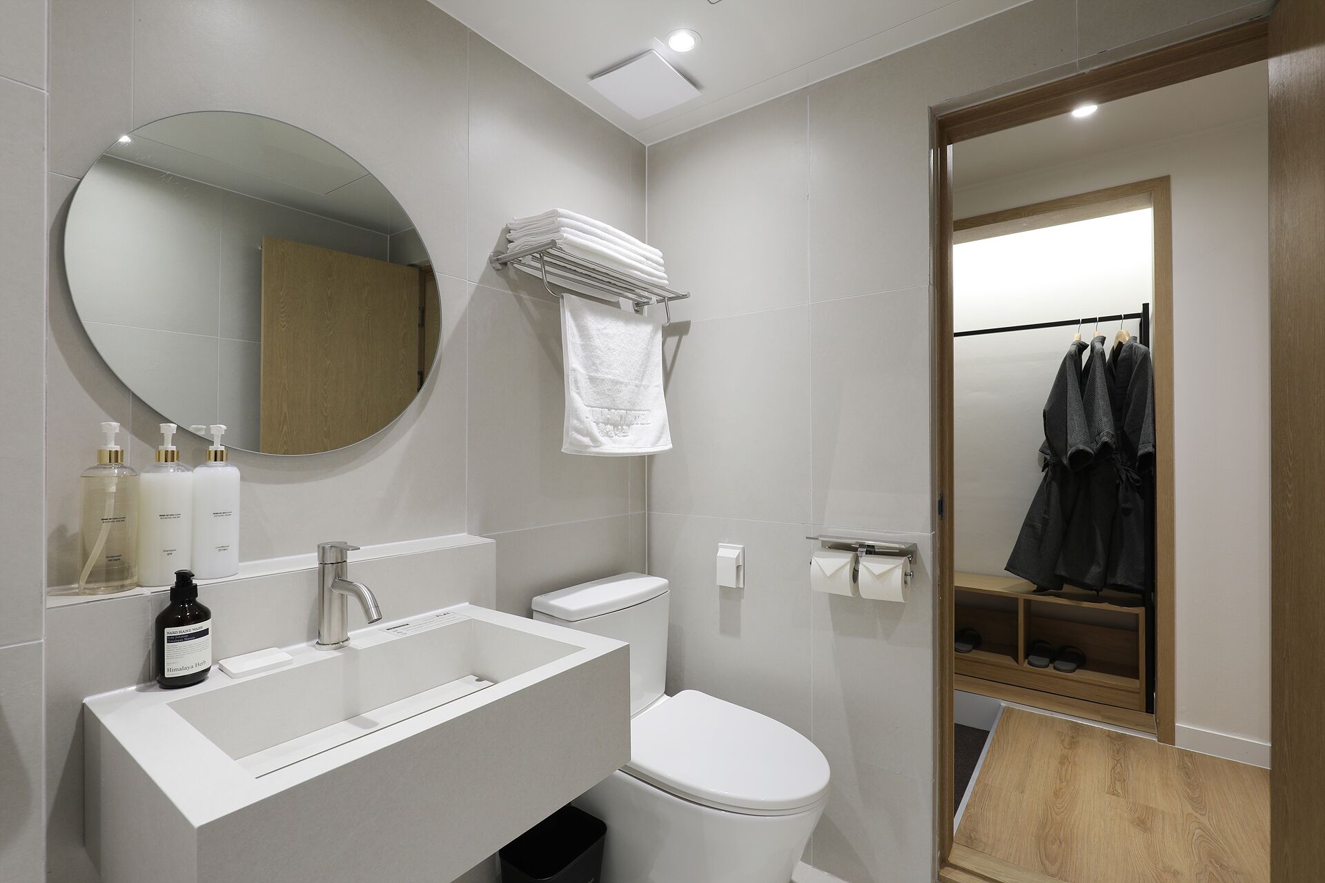 A modern bathroom with a white sink, toilet, and mirror. There are toiletries on the sink, a towel rack, and a robe rack with robes.