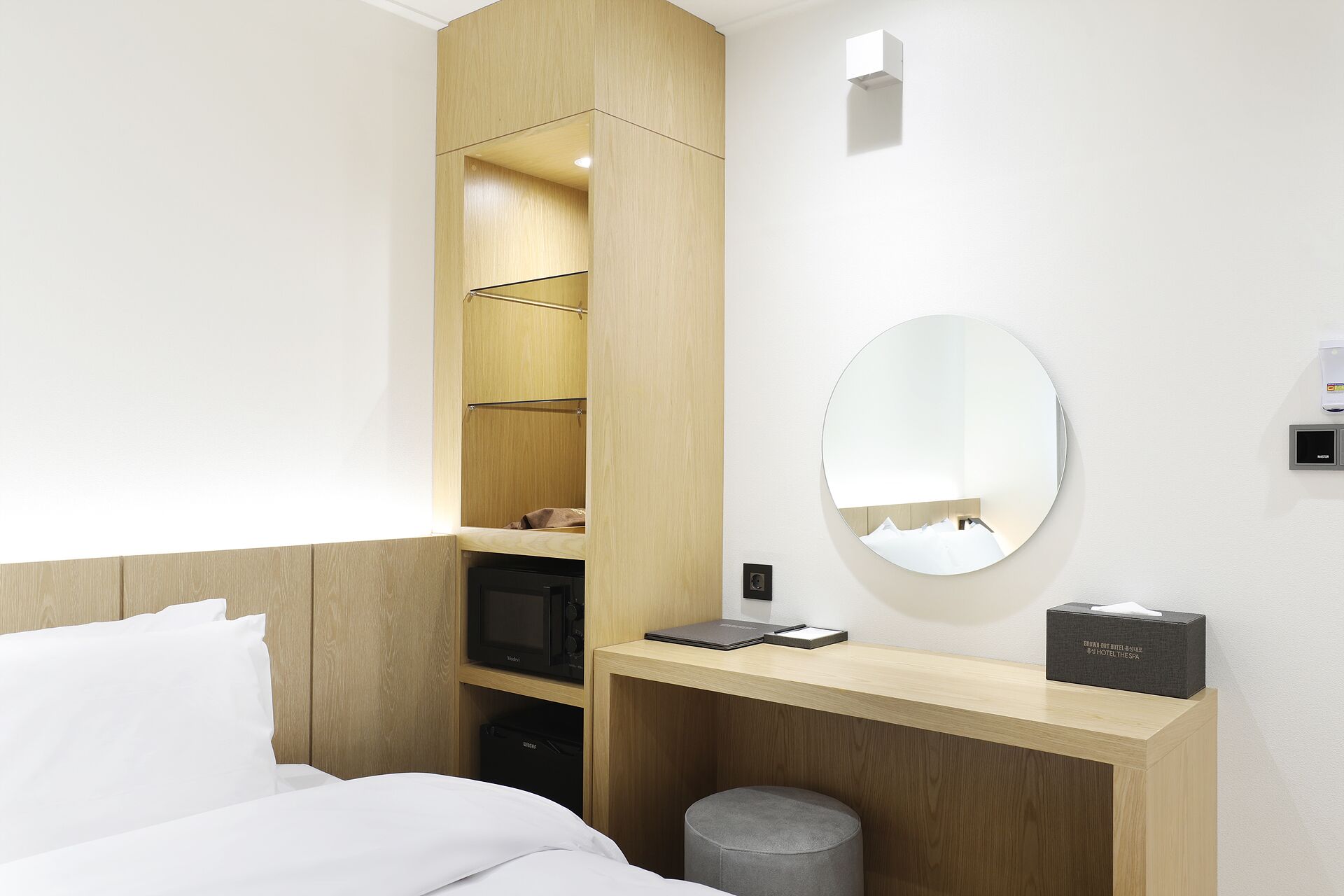 A hotel room with a bed, a wooden wardrobe, a desk with a mirror, a trash bin, and a small round mirror on the wall.