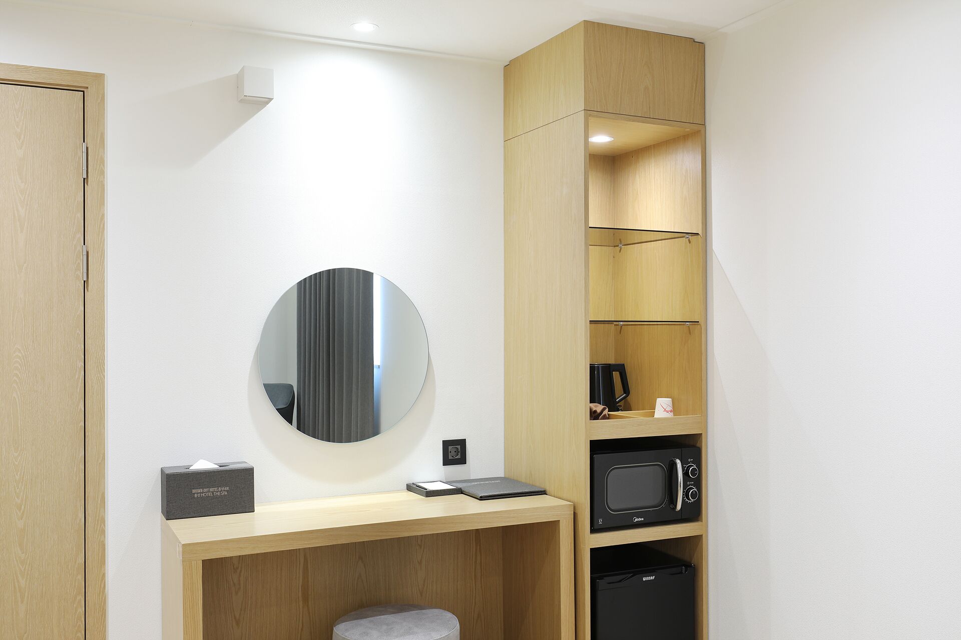 Una habitación de hotel compacta con un escritorio de madera, un espejo redondo, un microondas negro y un pequeño rincón de descanso.