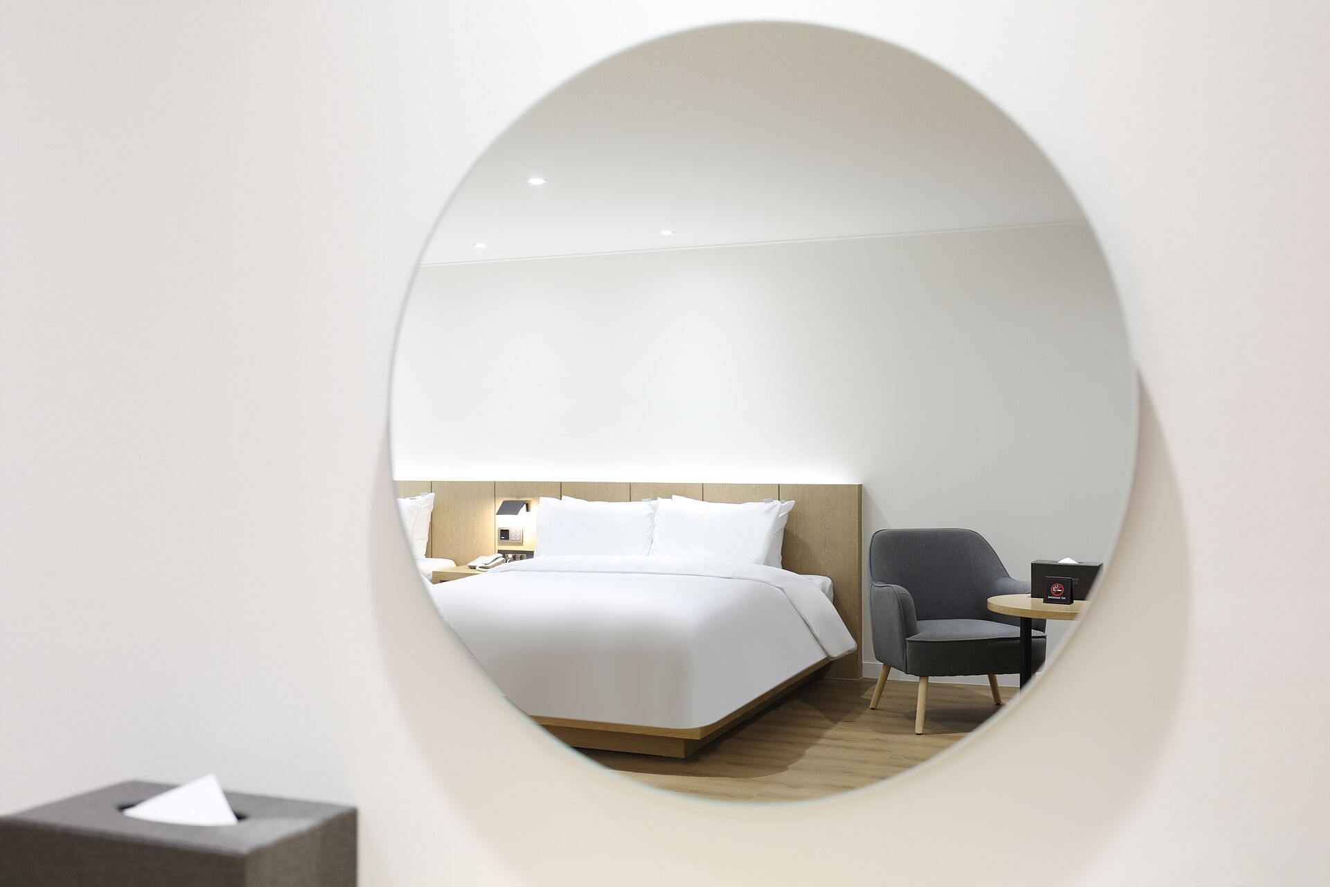 Ein modernes Hotelzimmer mit einem großen, ovalen Spiegel, der ein ordentlich bezogenes Bett, einen Nachttisch mit Lampe, einen Sessel und einen kleinen runden Tisch mit Telefon und Taschentuchbox widerspiegelt.