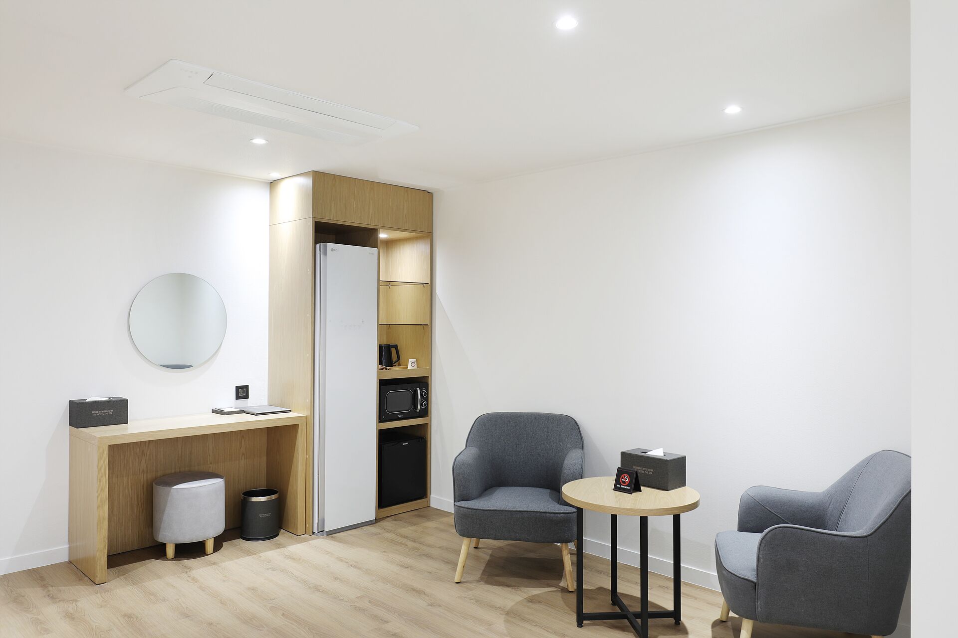 A modern, minimalist room with a wooden desk, two grey armchairs, a round mirror, and built-in shelving.