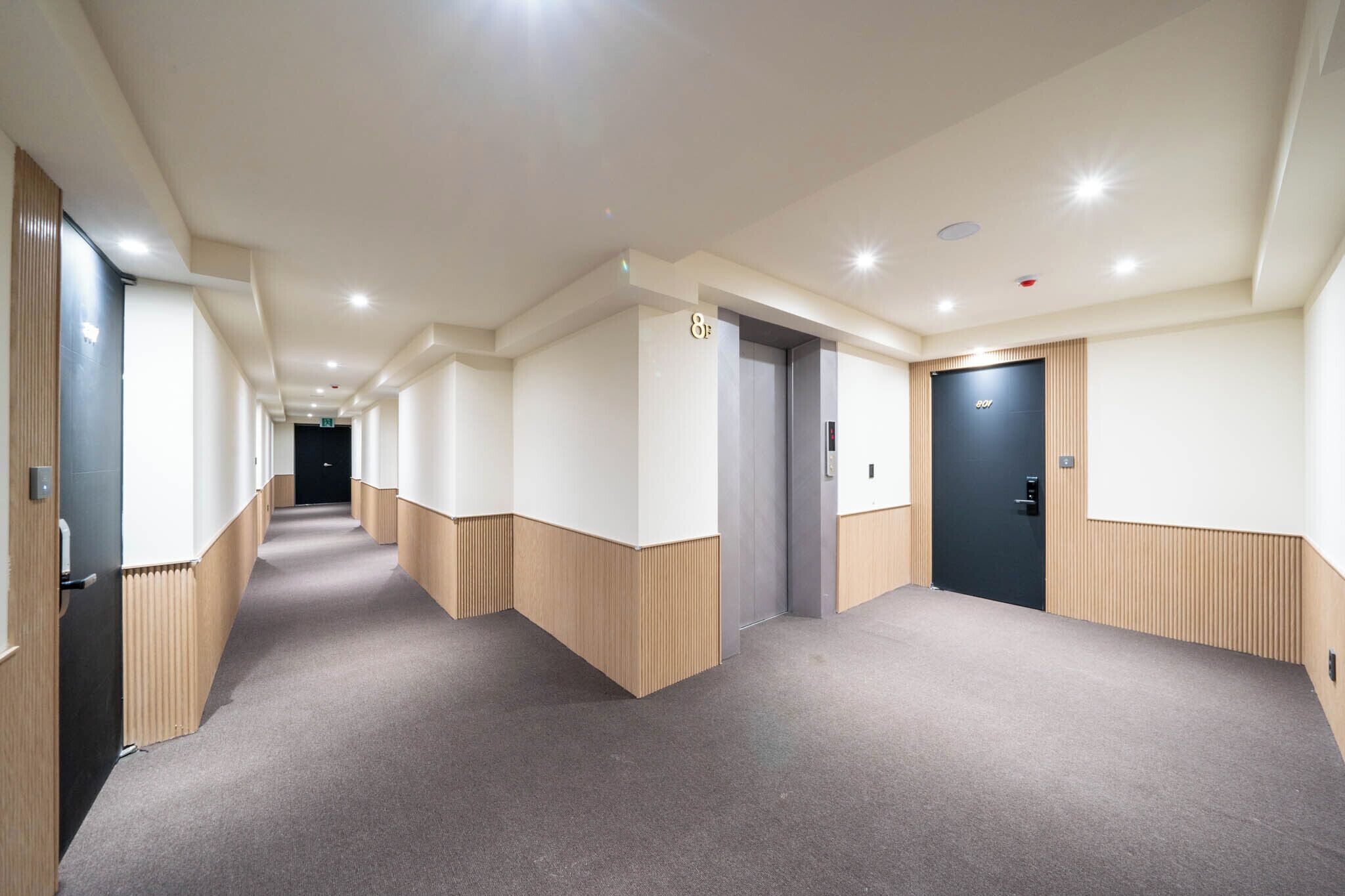 A modern hotel hallway with a carpeted floor, wooden paneling on the walls, and doors leading to rooms.