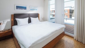 A hotel room with a large bed, wooden headboard, white bedding, a nightstand with a lamp, and a view of the city through a large window.