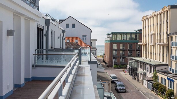A modern building with a balcony, a street with cars and pedestrians, and a view of the sea.