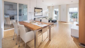 A modern apartment with a wooden dining table, white chairs, a kitchen with glass doors, a living area with a sofa, and a balcony with a view.