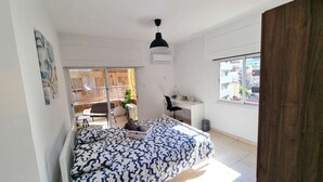 Un dormitorio con cama, escritorio, silla y ventana con vista a edificios.