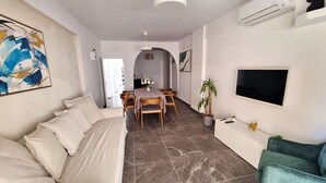 Una sala de estar moderna con un sofá, un comedor con mesa y sillas, un televisor montado en la pared y una planta en maceta.