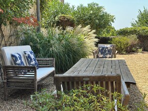 A wooden bench and a wooden table with a cushion, surrounded by greenery and a gravel path.