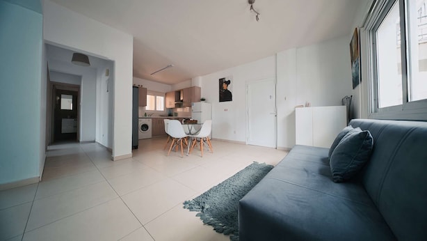 Una sala moderna con un sofá gris, un comedor con mesa y sillas, una cocina con armarios y electrodomésticos, y un pasillo que lleva a otra habitación.