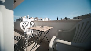 Un balcón con mesa y sillas, cielo despejado y edificios al fondo.